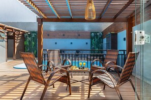 Um deck de madeira com cadeiras de vime e uma mesa, piscina ao fundo e guarda-corpo de vidro.