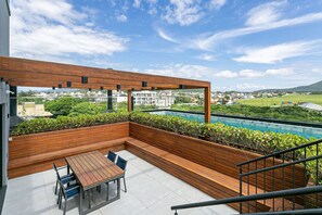A rooftop patio with a wooden deck, a table with chairs, and a view of a swimming pool and buildings in the distance. - Steps from the sea in Novo Campeche: high-end with pool (Florianópolis)