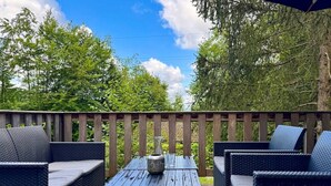 A deck with wicker furniture and a table, surrounded by trees and a wooden railing. - Cozy Durbuy Chalet With Fireplace (Rendeux)
