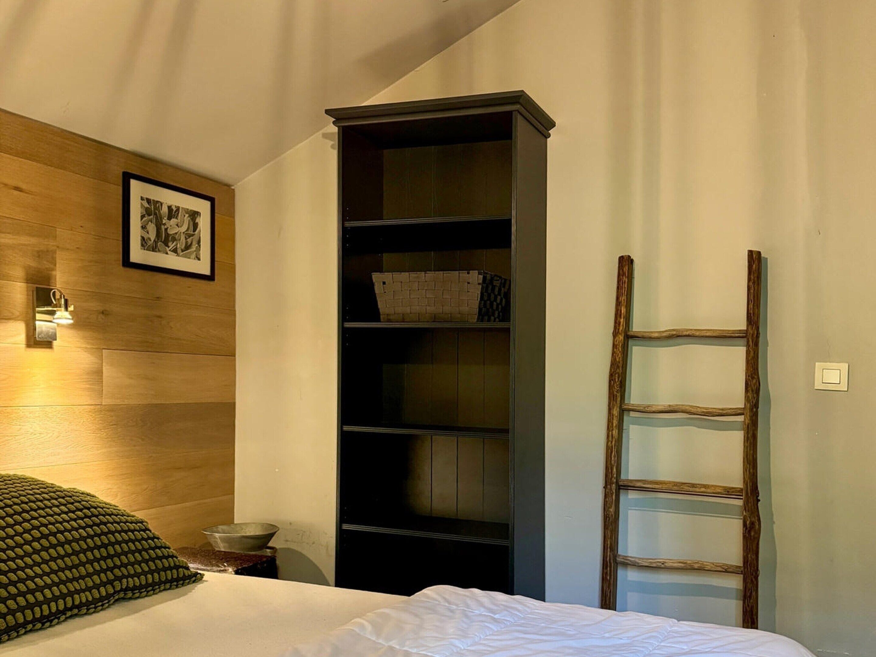 A bedroom with a bed, a wooden headboard, a black shelving unit, a ladder, and a framed picture on the wall.