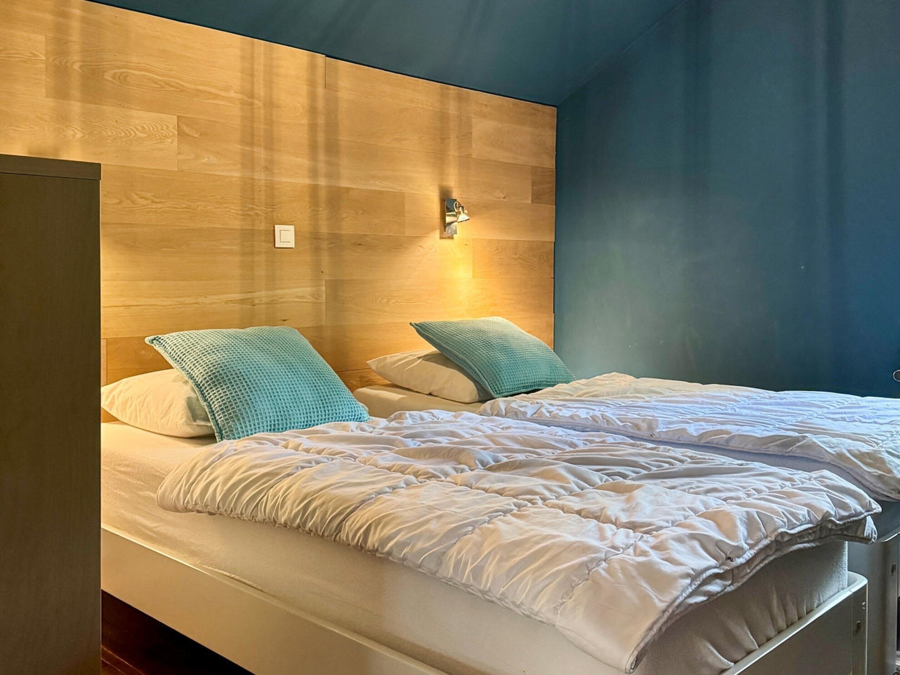 Une chambre avec deux lits, une tête de lit en bois, des coussins de couleur turquoise et un luminaire fixé au mur.