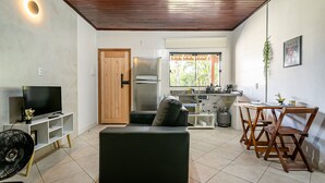 Uma sala de estar com sofá, TV e ventilador, uma cozinha com geladeira e uma área de jantar com cadeiras e mesa.