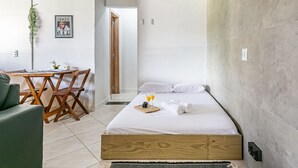 Quarto com cama de madeira, roupa de cama branca, uma mesinha com bandeja de suco de laranja e uma cadeira verde.
