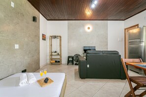 A hotel room with a bed, a sofa, a dining table, a refrigerator, and a fan. - Studio #03 Armação Beach Nature & Peace (Florianópolis)