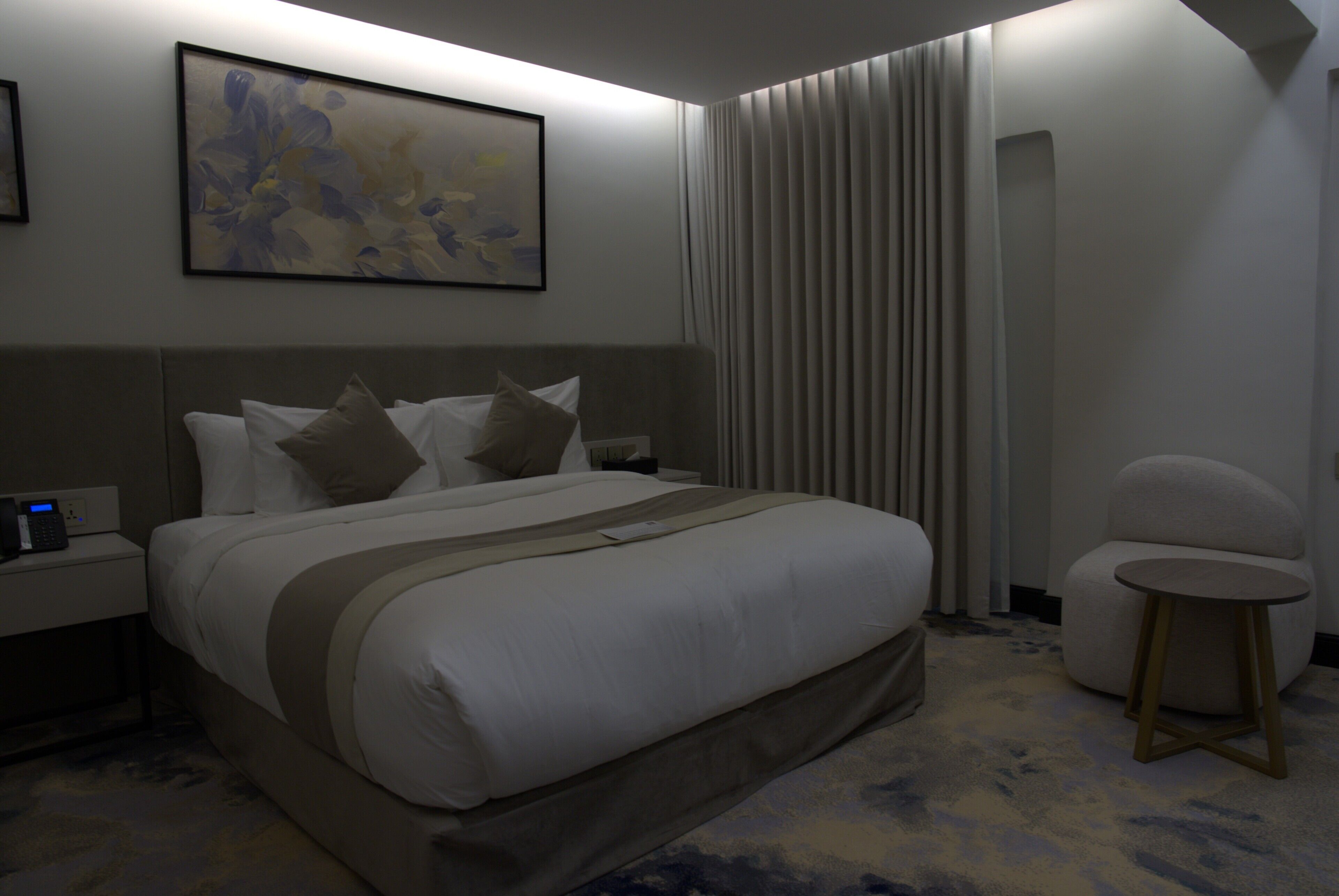 Una camera d'albergo con un letto grande, un comodino con il telefono, un tavolino rotondo e un quadro appeso al muro.