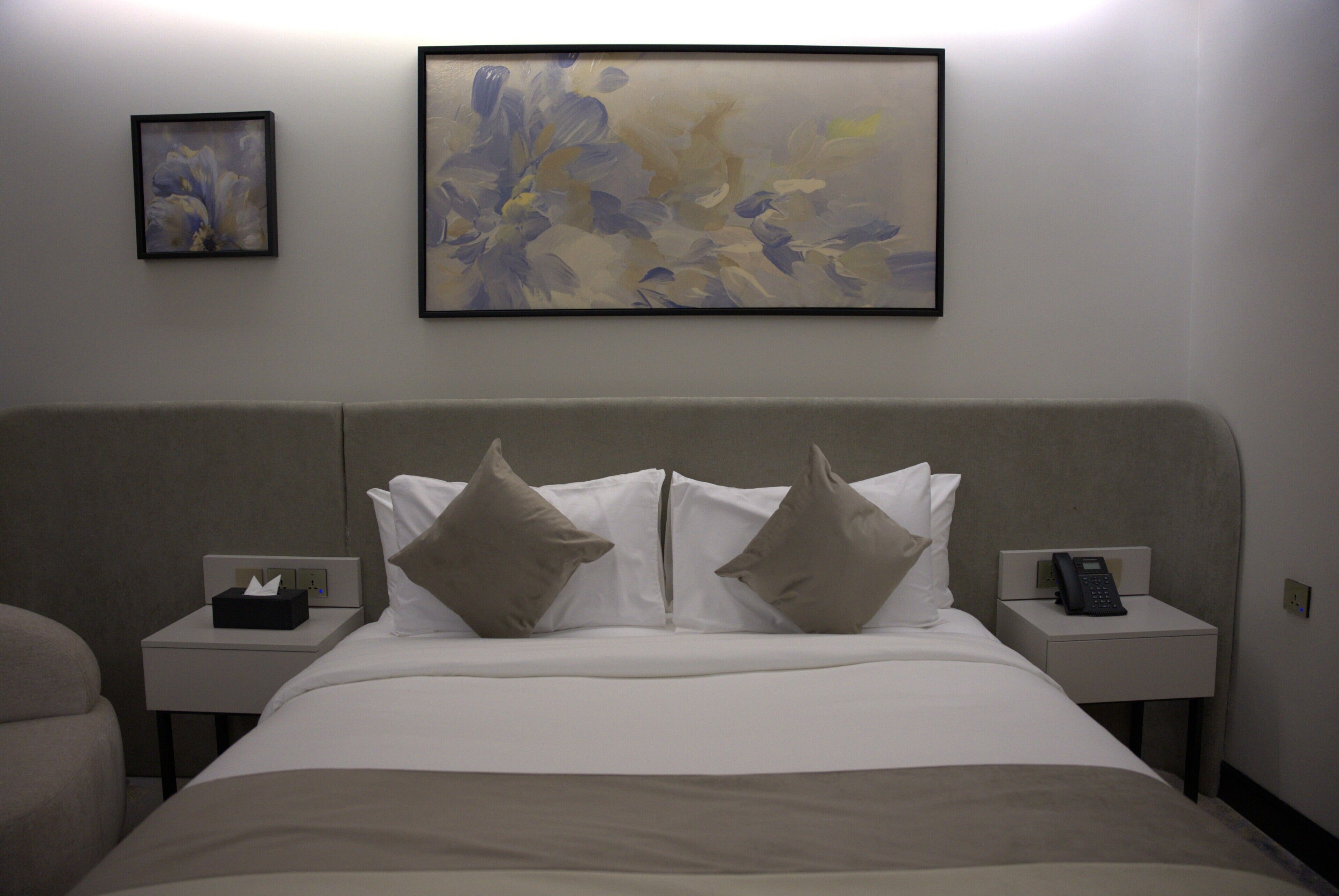 A hotel room with a bed, two bedside tables, a telephone, and a framed abstract painting on the wall.