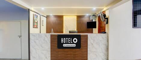 A hotel reception desk with a marble counter, wooden paneling, a flat-screen TV, and a sign indicating it's a company-serviced hotel.