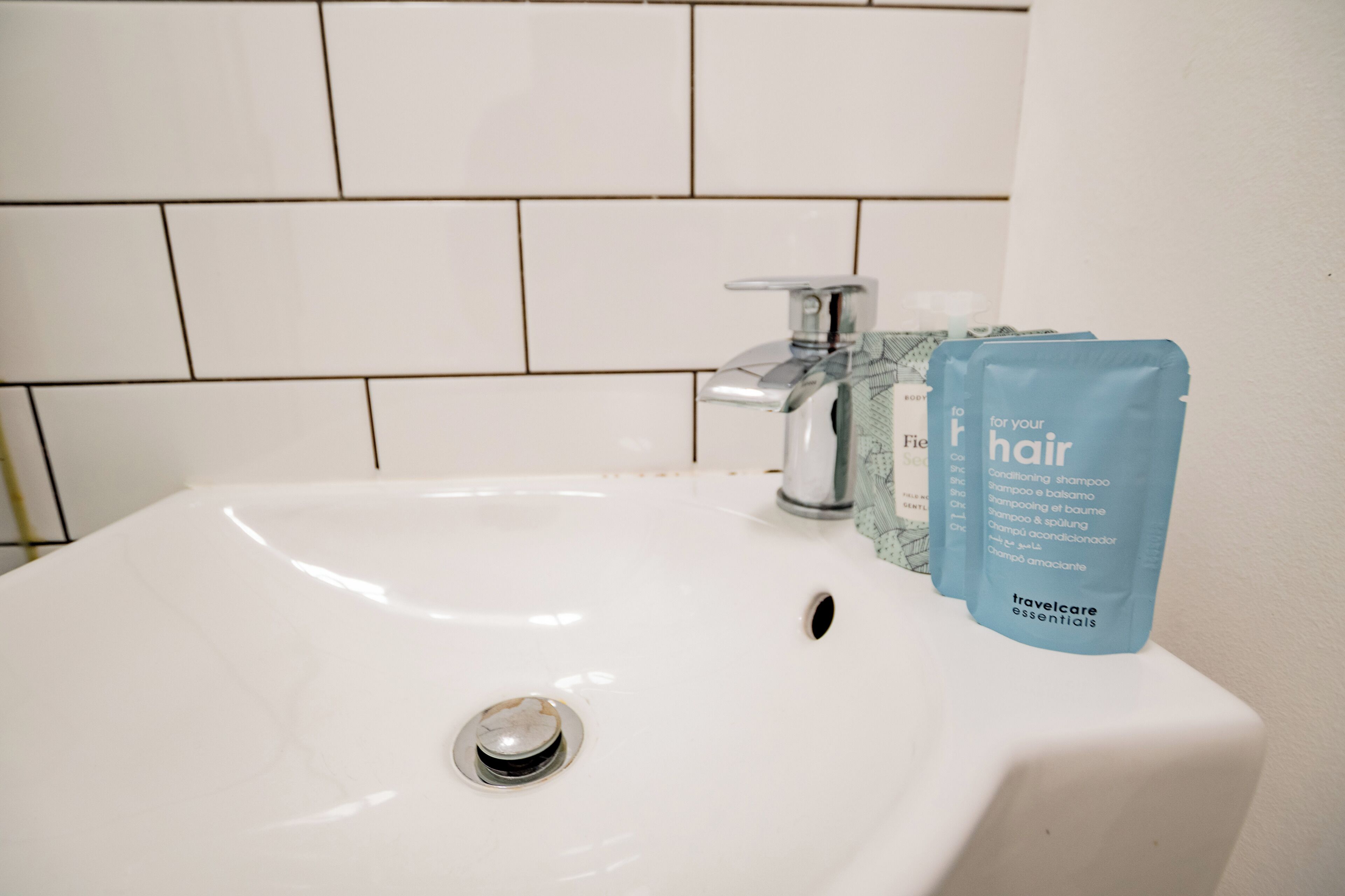 A white bathroom sink with a chrome faucet, a blue toiletry bag labeled 'travelcare essentials,' and a tiled wall in the background.