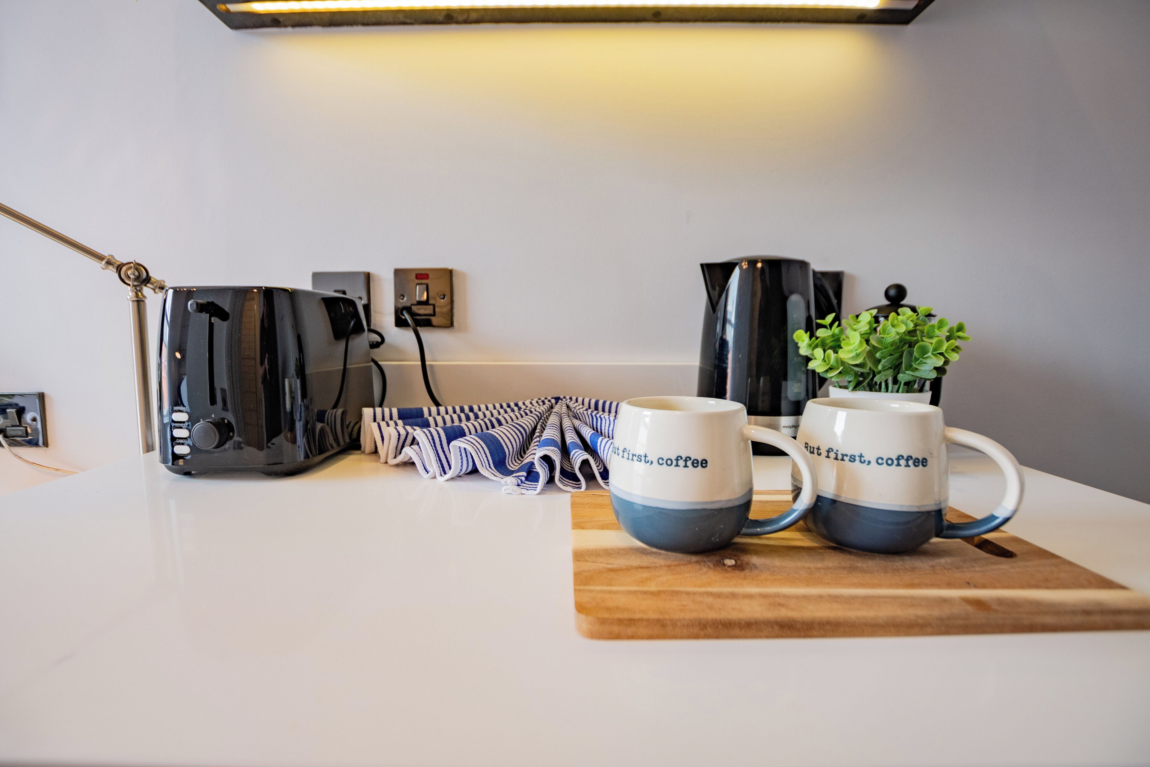 Una encimera de cocina con una cafetera, dos tazas, una tabla de cortar y un enchufe.