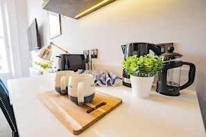 Una cocina con cafetera, dos tazas, tabla de cortar, planta en maceta y un paño a rayas.