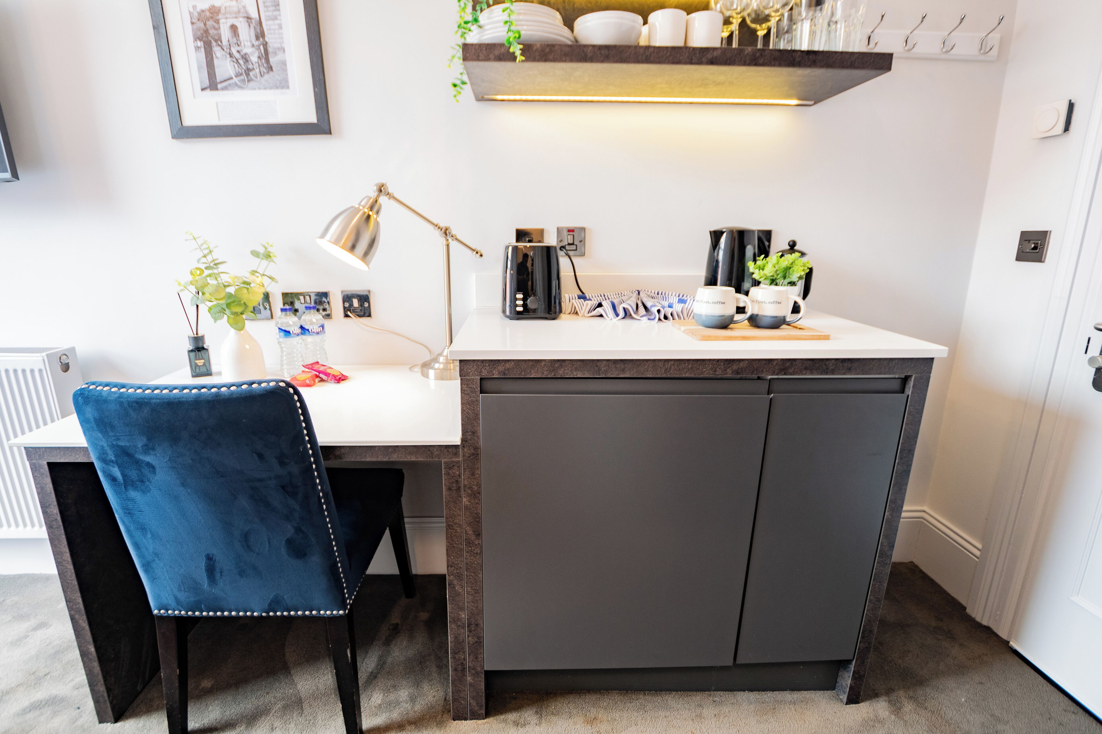 Une cuisine de taille réduite avec un plan de travail blanc, un meuble sombre, une chaise bleue, une lampe de bureau et une étagère contenant de la vaisselle et une plante.