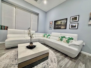 Un salon moderne avec un canapĂ© dâangle blanc, une table basse et une grande fenĂȘtre Ă©quipĂ©e de stores.