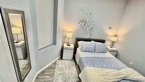 A bedroom with a bed, bedside lamps, a mirror, a window with blinds, and a wall art piece.