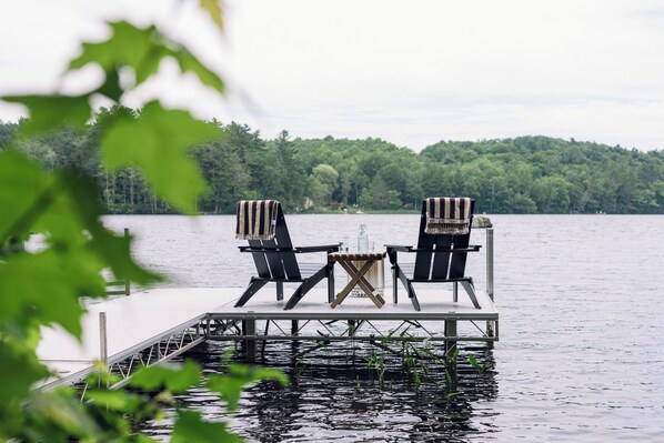 A dock with two Adirondack chairs and a small table, overlooking a calm lake with a forested shoreline.