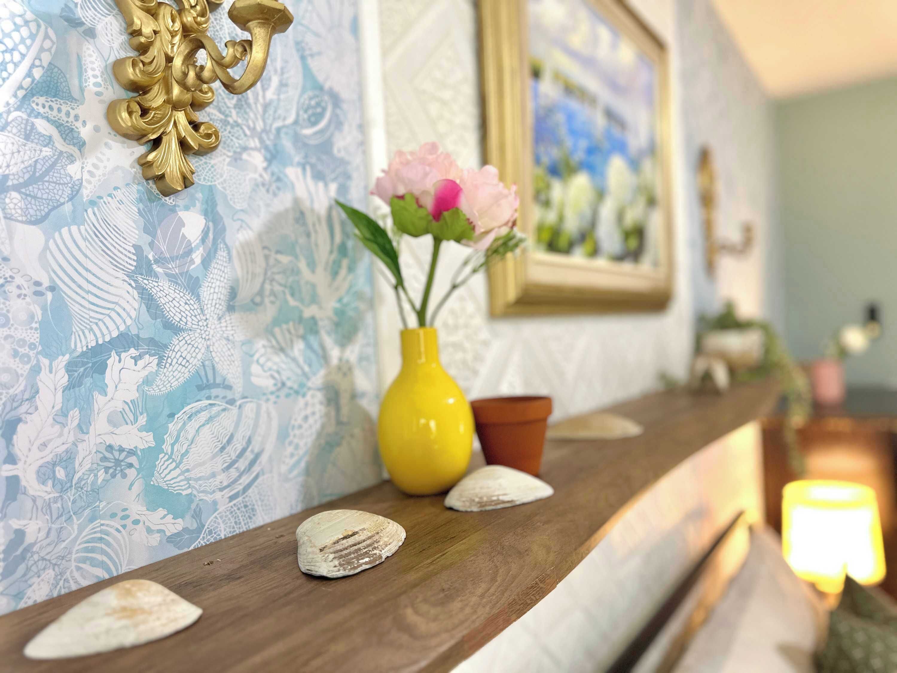 A wooden shelf with decorative shells, a yellow vase with flowers, and a framed picture on a wall with a blue and white pattern.