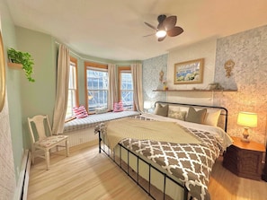 A bedroom with a bed, a window seat, a chair, a bedside table, a lamp, and a ceiling fan.