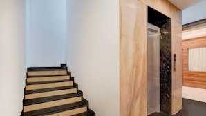 Uma escadaria moderna com degraus pretos e soleiras bege, que leva a um elevador com acabamento em mármore.
