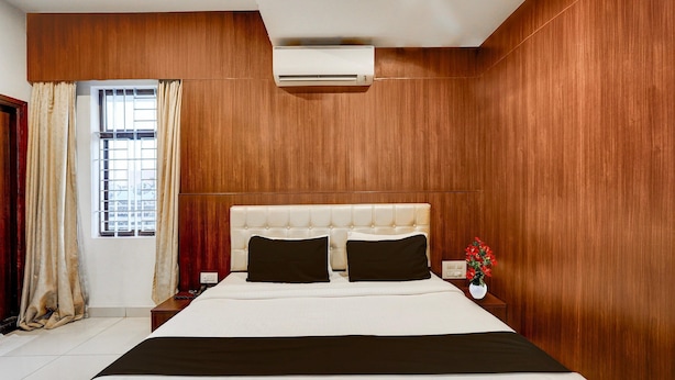 Quarto de hotel com parede de painel de madeira, cama com roupa de cama branca, duas almofadas pretas, mesa de cabeceira com um vaso de flores e um aparelho de ar condicionado no teto.