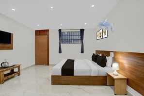 Una habitación de hotel con cama, mesita de noche, lámpara, ventilador en la pared, ventana con cortinas y televisor.