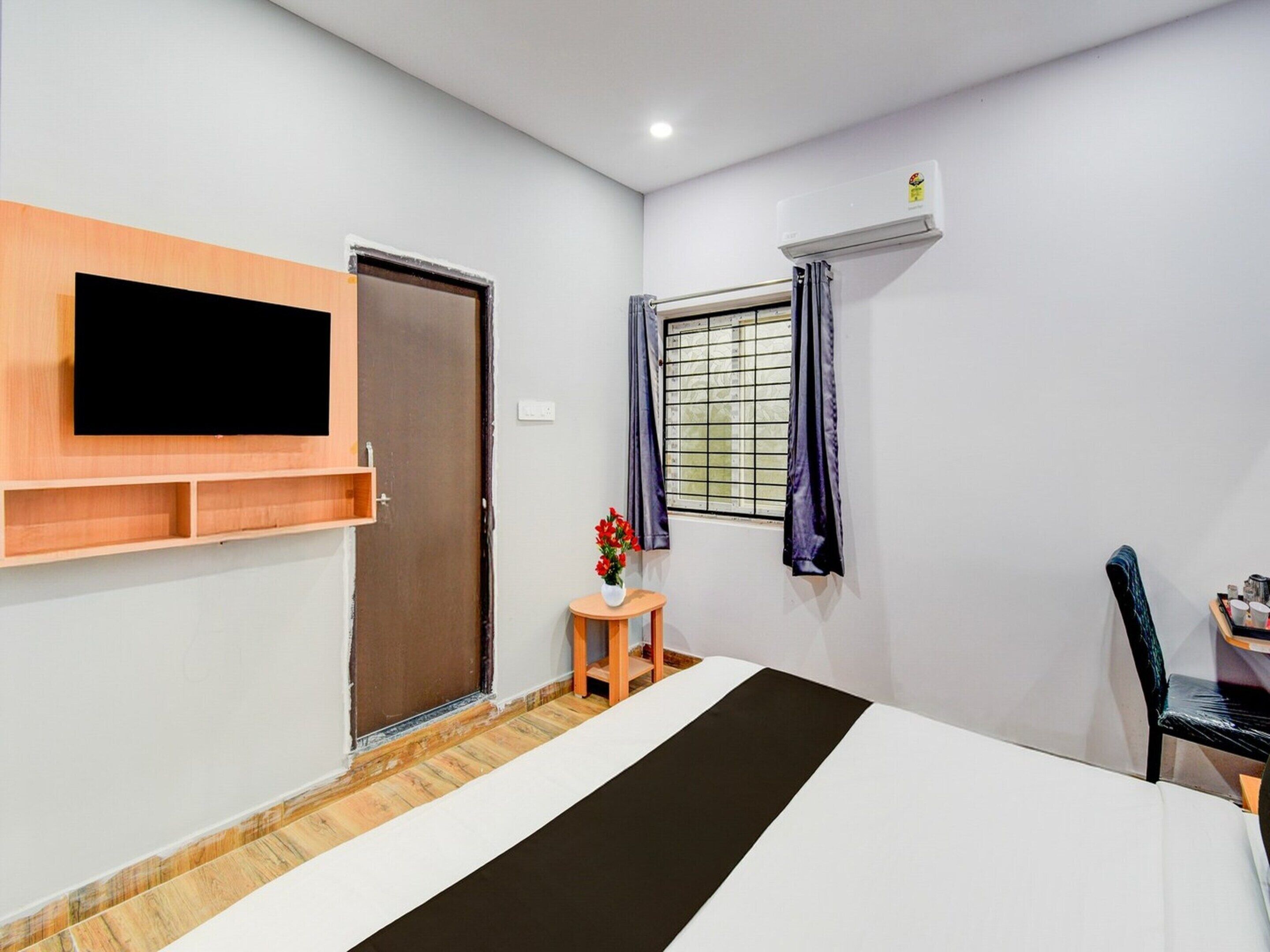 A hotel room with a bed, a TV mounted on the wall, a window with curtains, a small table with a vase, and an air conditioning unit.