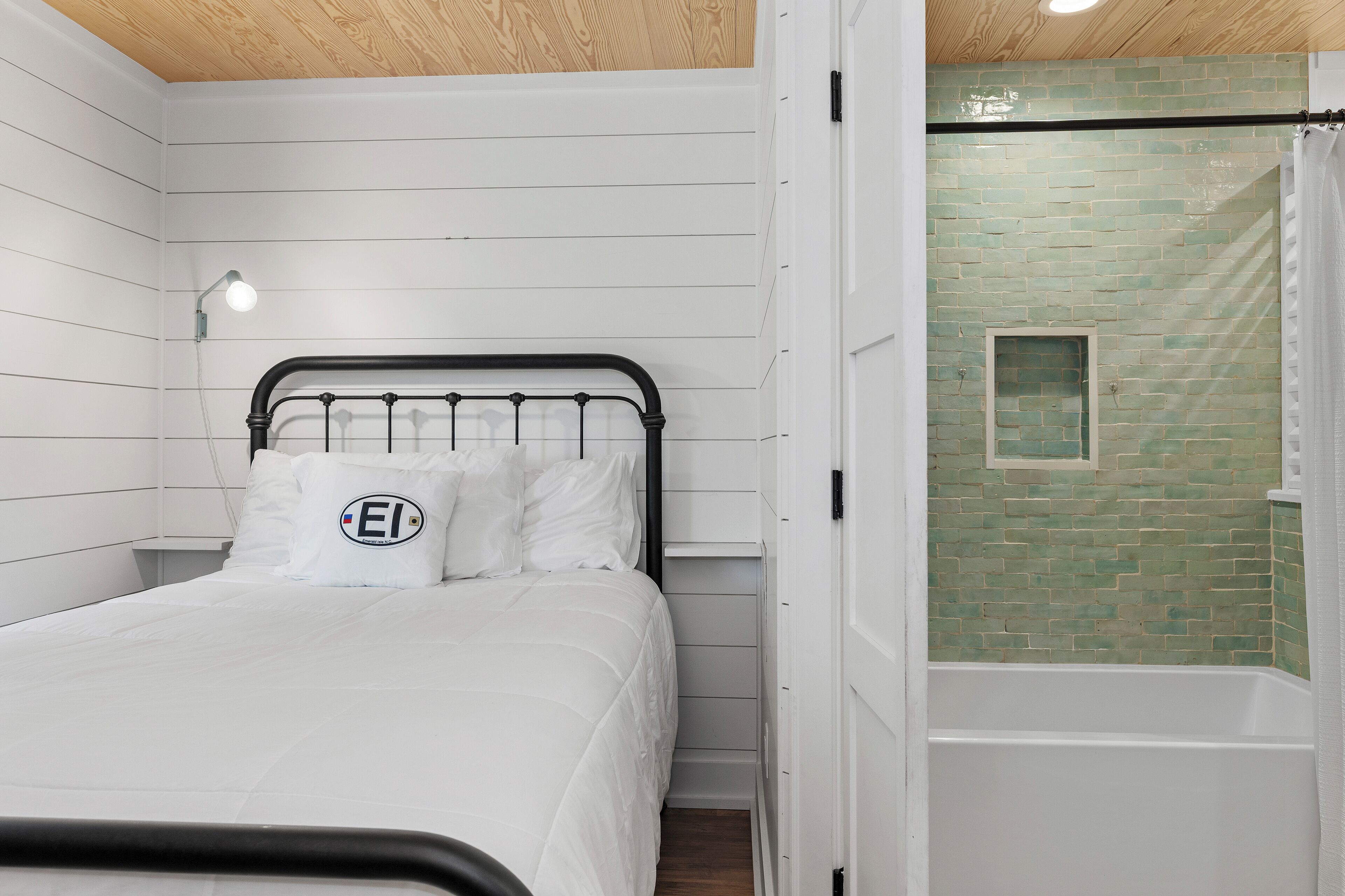 A bedroom with a bed, a nightstand, a wall-mounted light, and a bathroom with a shower and a green-tiled wall.