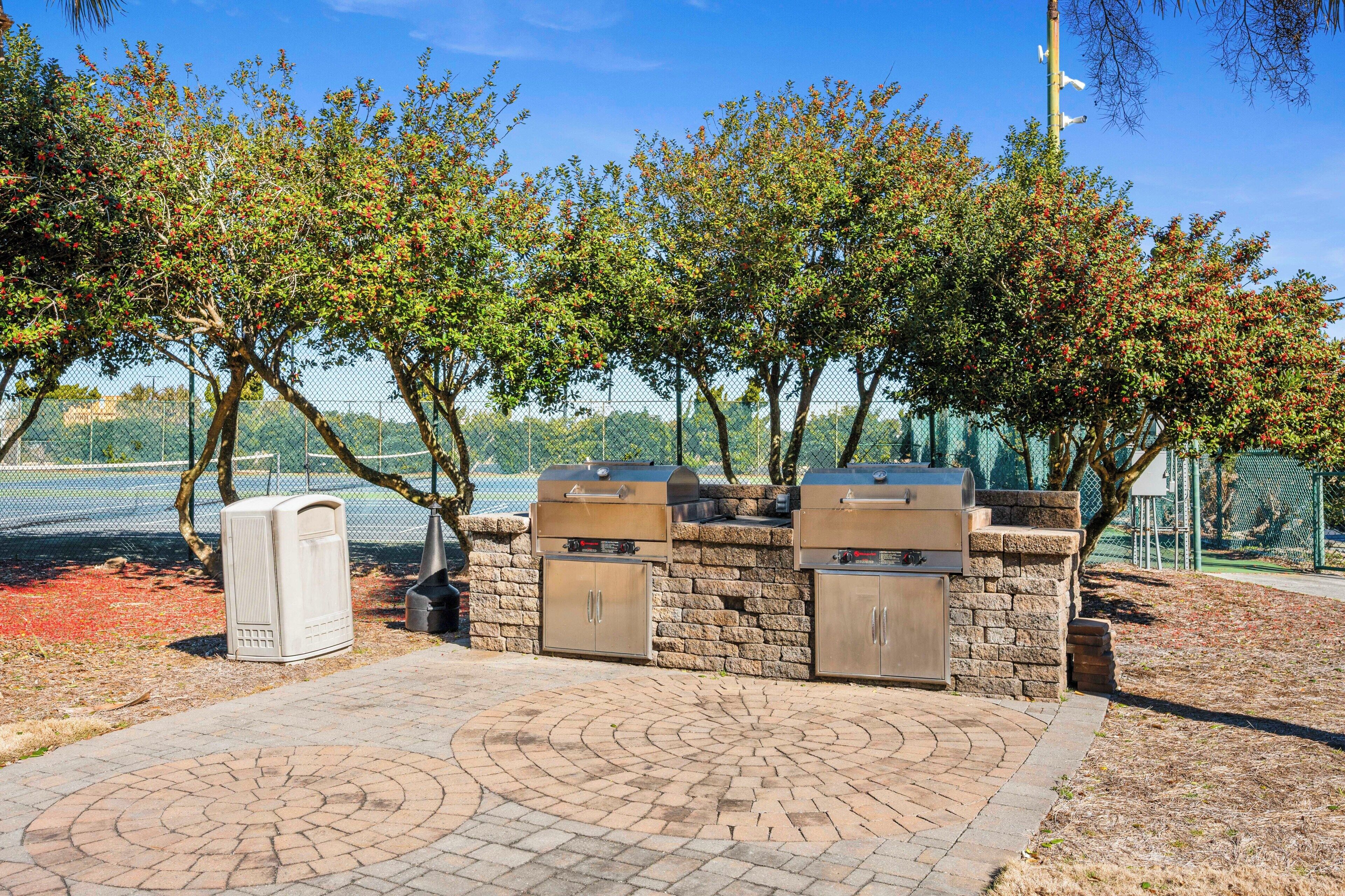 An outdoor barbecue area with stone walls and grills, surrounded by trees and a fence.