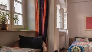 Ein Zimmer mit Bett, Fenster, Sessel, Tisch und einer Topfpflanze.