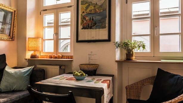 Ein gemütliches Zimmer mit Esstisch, Stühlen, einer Couch, einer Lampe, einem gerahmten Bild und einem Fenster mit Landschaftsblick.