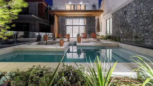 Un área de piscina moderna al aire libre con pared de piedra, sillas de descanso y una pérgola de madera.