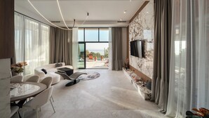 Una sala de estar moderna con sofá, sillas, una mesa de comedor, televisor montado en una pared de mármol y vista al exterior.