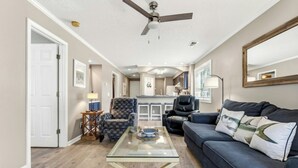 A living room with a glass coffee table, a dark sofa, a ceiling fan, and a kitchen in the background. - Tilghman Shores H1- 2 Bedroom- Short Walk To Beach (North Myrtle Beach)