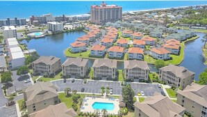 A residential area with a central swimming pool, surrounded by single-family homes with red roofs, and a large hotel building in the background. - Tilghman Shores H1- 2 Bedroom- Short Walk To Beach (North Myrtle Beach)