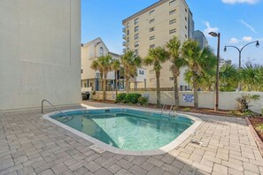Ein Swimmingpool mit Backstein-Terrasse, umgeben von Palmen und einem mehrstöckigen Gebäude im Hintergrund.