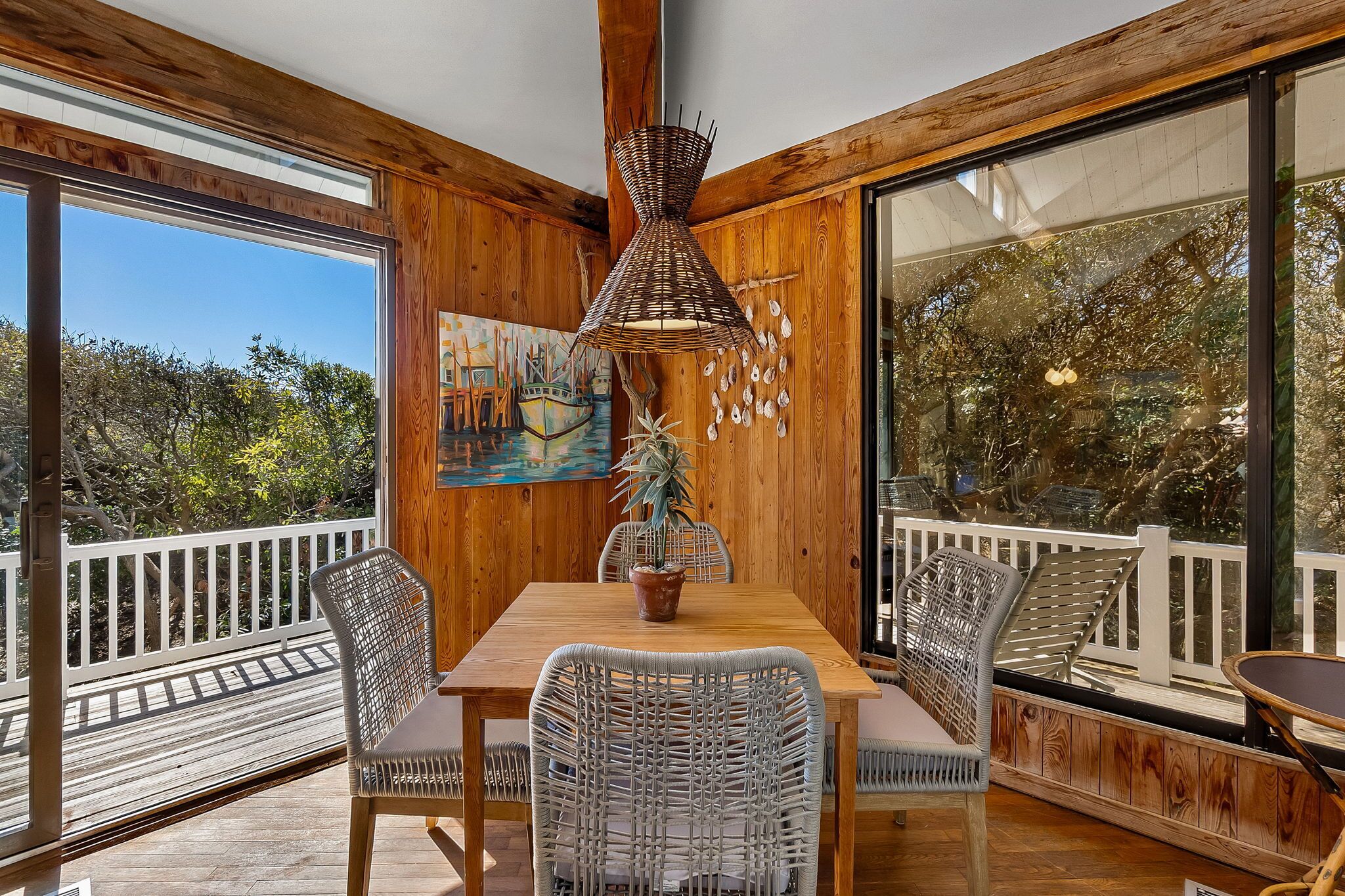 A wooden interior with a dining table, wicker chairs, a hanging light fixture, and a large window with a view of trees.