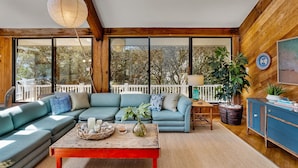 A modern living room with a sectional sofa, a coffee table, and a dining area. - Oceanfront Treehouse Retreat | Private Beach Walkw (Pine Knoll Shores)