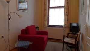 Un piccolo soggiorno con un divano rosso, una TV, una lampada e una finestra con tende.