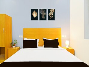 A hotel room with a wooden headboard, a bed with white bedding, black and white pillows, a nightstand with a lamp, and three framed wall art pieces. - Deluxemedipally (Hyderabad)