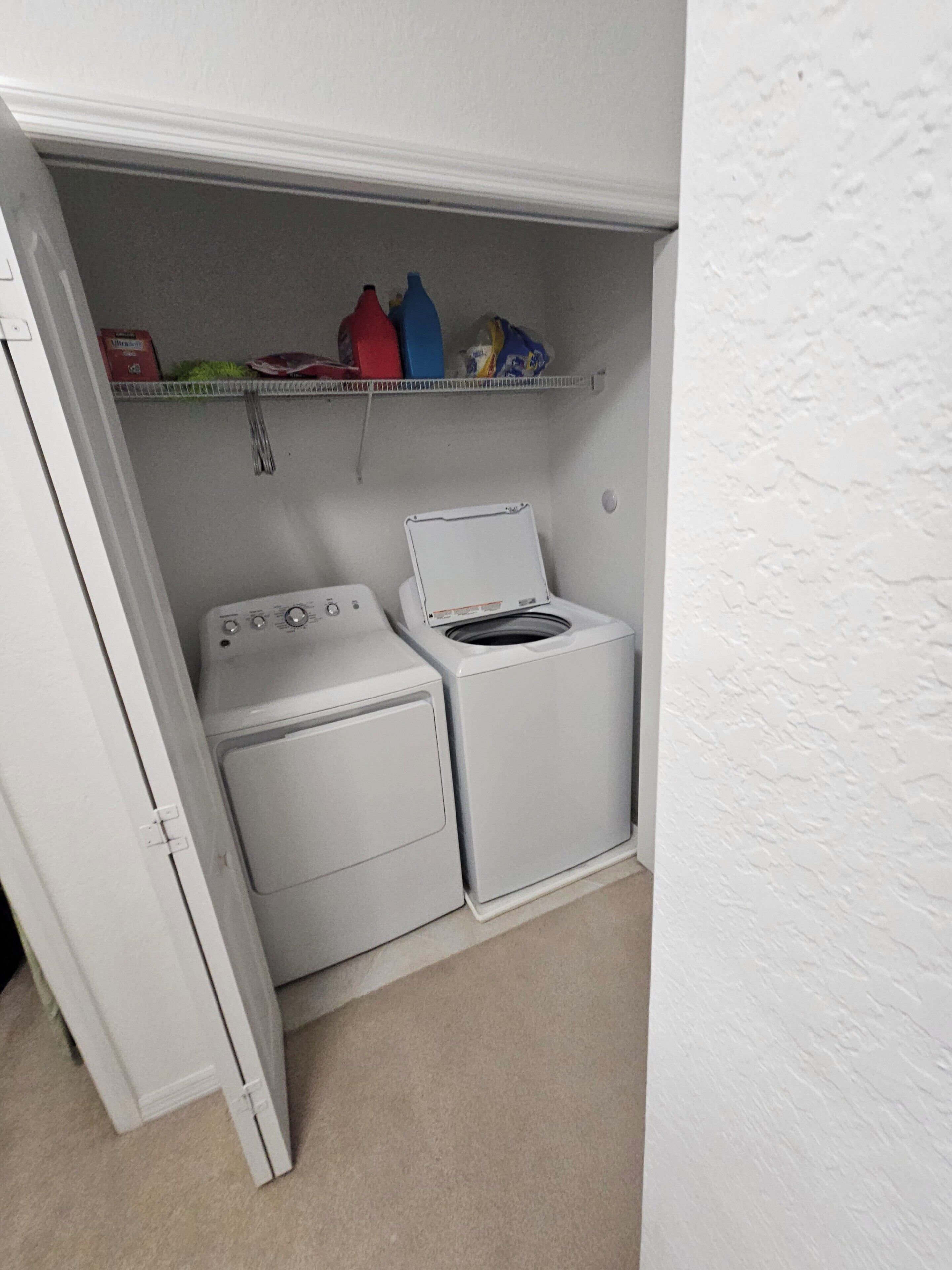 A laundry room with a washing machine and dryer, a shelf with cleaning supplies, and a door leading to another room.