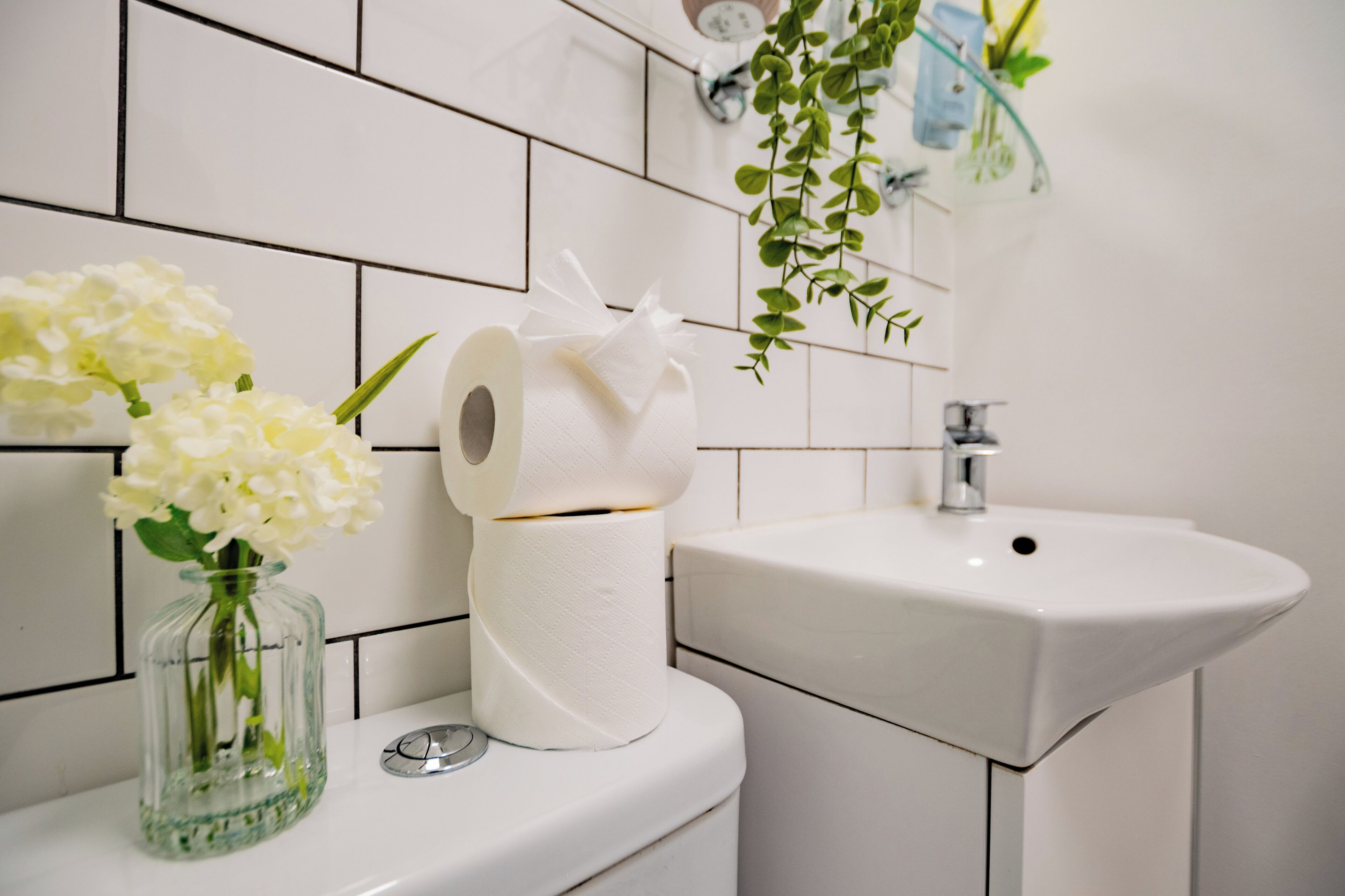 A bathroom with a toilet, a sink, a glass vase with flowers, and a roll of toilet paper.