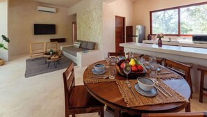 A dining area with a round table set for four, a kitchen with a large window, and a living area with a sofa and TV. - Holistika 3BR Jungle Escape with Plunge Pool (Tulum)