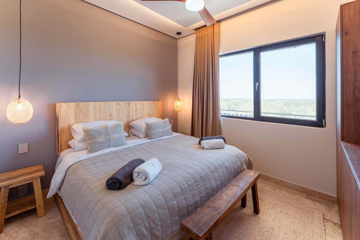 Un dormitorio con una cama grande, cabecera de madera, una banca, una mesita de noche y una ventana con cortinas.