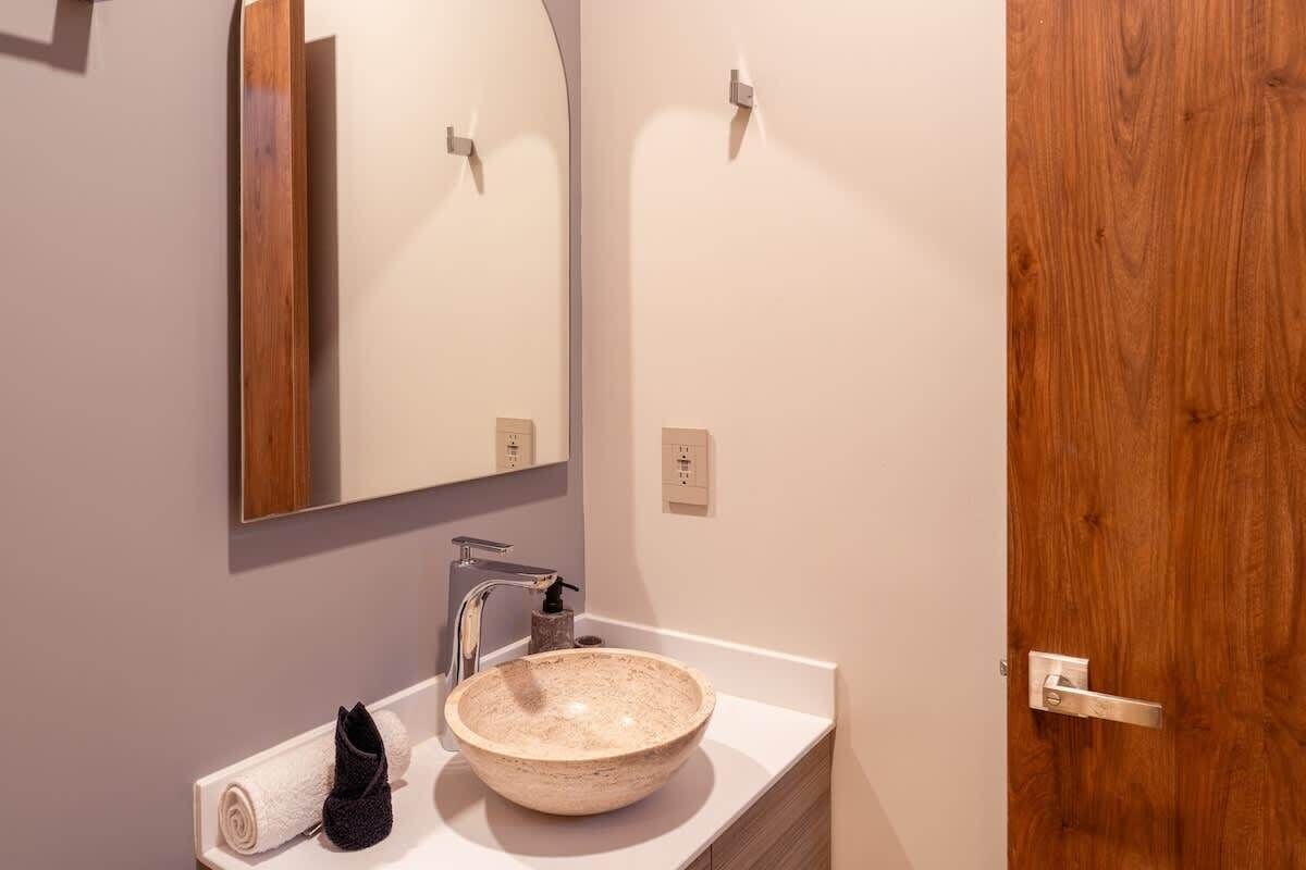 Une salle de bain avec une porte en bois, un lavabo blanc avec un récipient beige, un miroir au-dessus du lavabo et une serviette posée sur le comptoir.