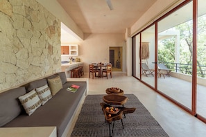 A modern interior with a stone wall, a sofa with cushions, a dining area, and a balcony with outdoor furniture. - Ahal Luxe 3BR Penthouse – Rooftop & Plunge Pools (Tulum)