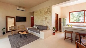 Una sala de estar moderna con sofá, televisor, pared de piedra, zona de cocina con refrigerador y ventana.