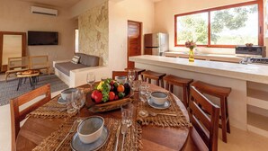 Un comedor con una mesa redonda preparada para cuatro, una cocina con refrigerador y microondas, y un gran ventanal que proporciona luz natural.