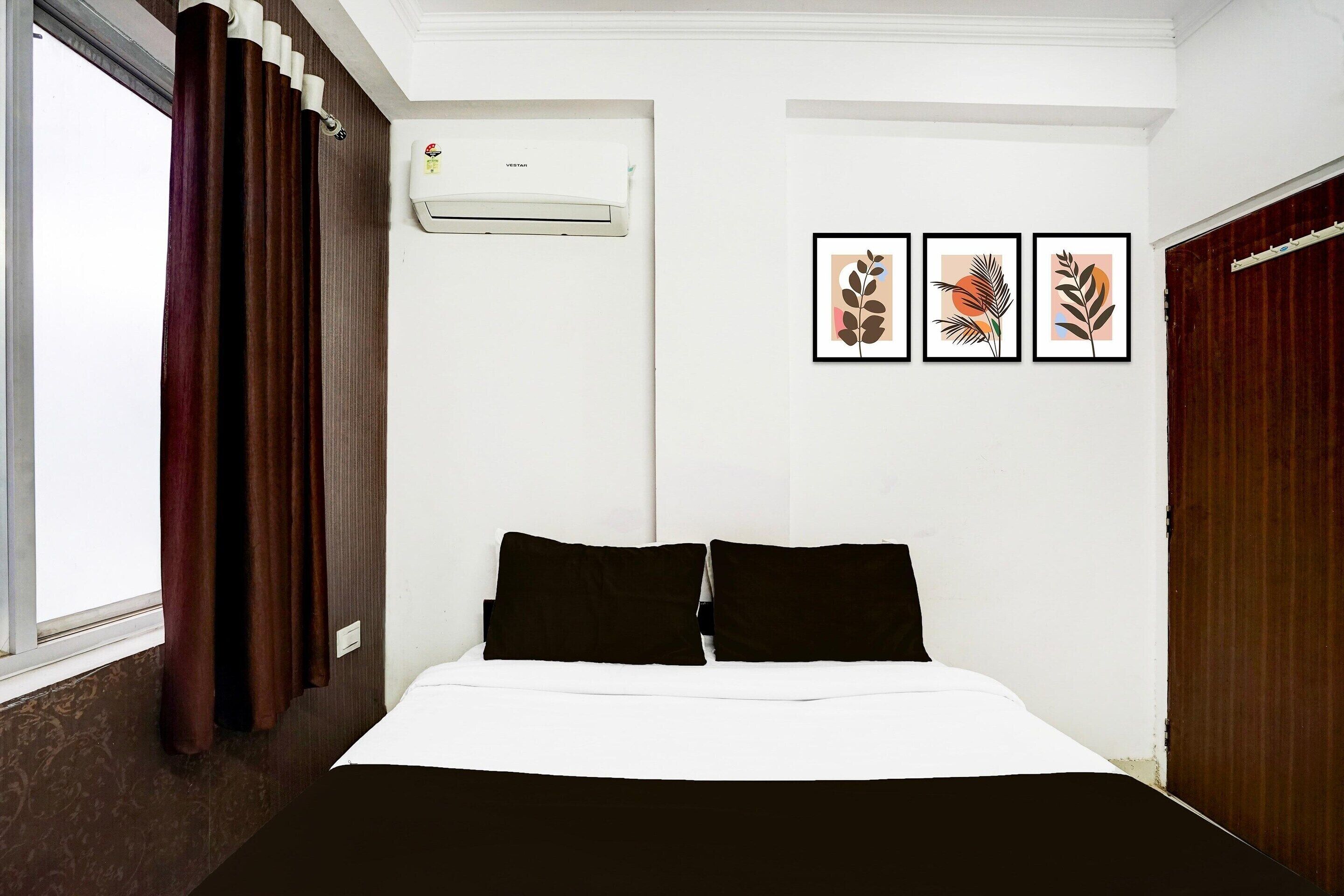 Habitación de hotel con cama, cortinas marrones, unidad de aire acondicionado y tres láminas con motivos vegetales en la pared.