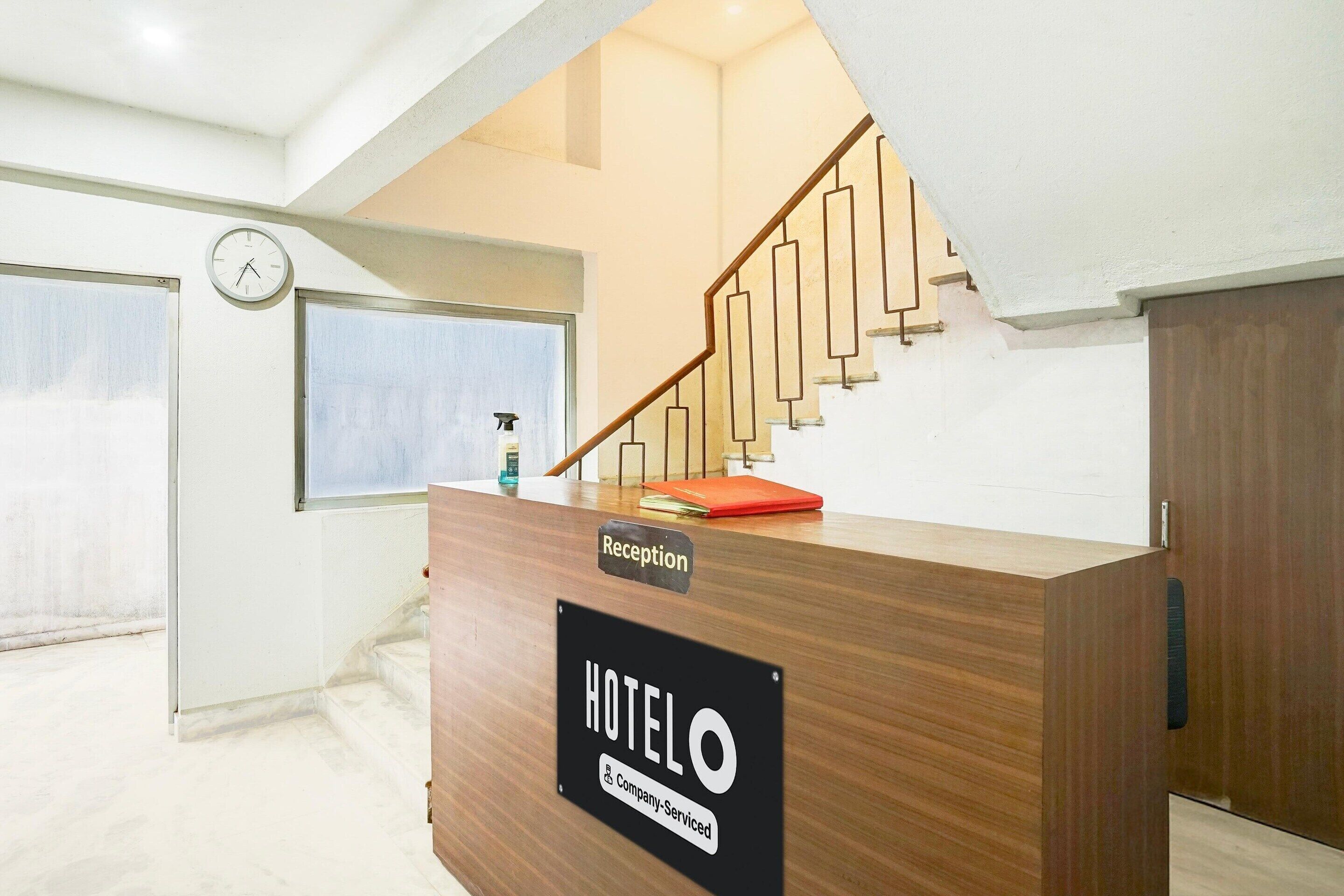 A reception desk labeled 'HOTELO' with a clock above it, a staircase with a wooden handrail, and a window with blinds.
