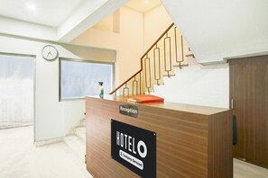 Vastaanottotiski, jossa on kyltti »HOTELO», sen yläpuolella kello, puinen kaide varustettu portaikko ja ikkuna, jossa on sälekaihtimet.