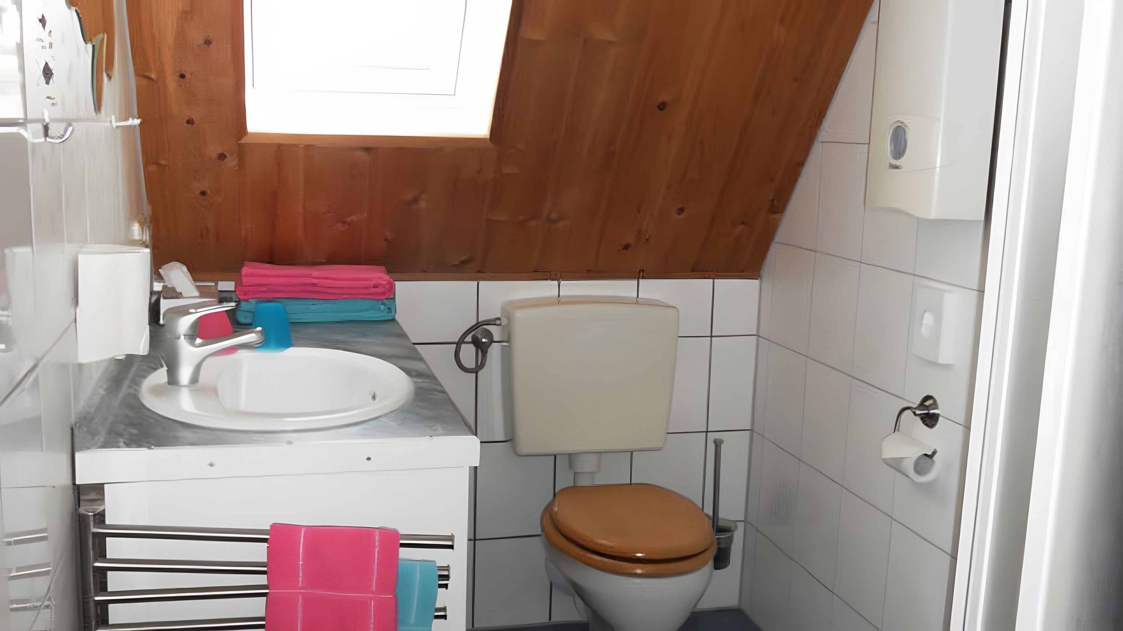 Een kleine badkamer met een houten plafond, een witte wastafel, een toilet en een raam.