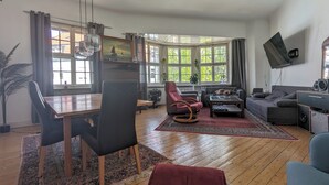 A spacious living room with a dining area, large windows, a mounted TV, and a cozy seating arrangement. - Cozy old building with sauna and view (Wuppertal)